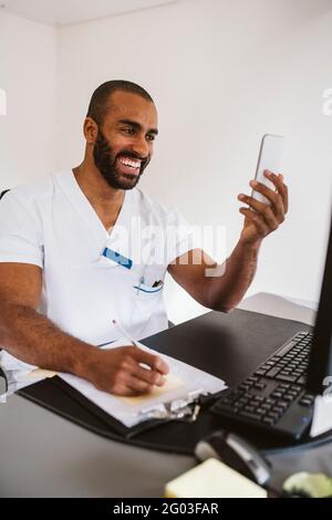 Un médecin homme joyeux consultant via un smartphone tout en écrivant à son bureau Banque D'Images