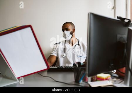Un médecin portant un masque facial de protection lisant des dossiers médicaux lorsqu'il est assis à son bureau Banque D'Images