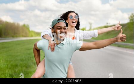 Couple multiracial en randonnée sur la route, en arrêtant la voiture avec le pouce vers le haut, en voyageant en auto-stop pendant les vacances d'été Banque D'Images