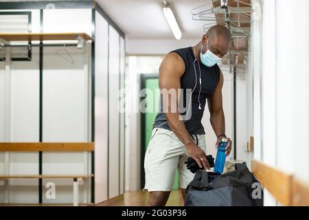 Un homme noir se prépare dans le vestiaire de la salle de gym. Il porte un masque médical. Nouveau concept normal. Banque D'Images