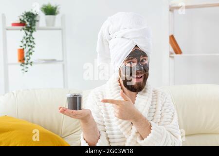 Homme barbu s'amusant avec un masque cosmétique sur son visage en argile noire. Hommes soins de la peau, humour et spa à la maison concept Banque D'Images