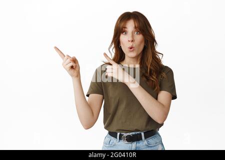 Curieuse fille poser la question sur la publicité, dites wow, regardant stupéfié à l'appareil photo, pointant à gauche à l'offre promotionnelle intéressante, des réductions dans le magasin Banque D'Images