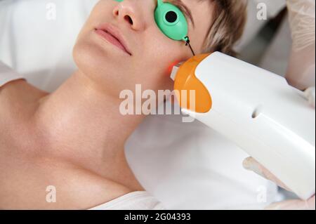 Vue partielle d'une jeune femme avec des lunettes de protection UV recevant un traitement laser avec un appareil laser moderne. Gros plan Banque D'Images