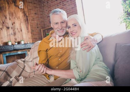 Photo de l'heureux positif vieux couple mari et femme assis canapé sourire câlin tenir les mains à l'intérieur à la maison week-end Banque D'Images