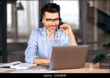 Confiant et amical jeune homme d'affaires caucasien, indépendant ou consultant portant des lunettes et des écouteurs, assis au bureau, utilisant un ordinateur portable, bavardant avec le client en ligne, tapant sur le clavier, souriant Banque D'Images