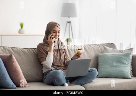 La femme arabe dans le hijab parle au téléphone, tient une carte de crédit et utilise un ordinateur portable Banque D'Images
