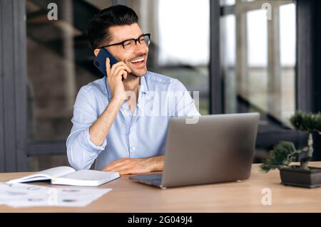 Conversation sur téléphone portable. Joyeux indépendant ou Manager caucasien parlant sur un smartphone tout en travaillant sur un ordinateur portable, un homme positif fait une conversation téléphonique agréable, regarde loin, sourit amical Banque D'Images