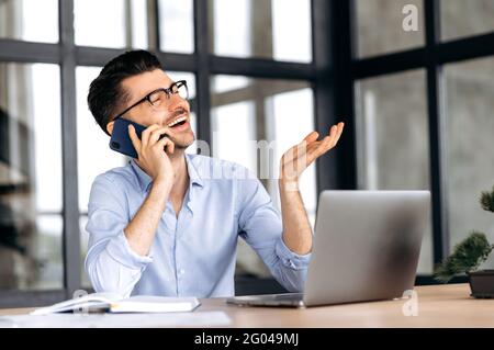 Communication par téléphone portable. Sympathique, joyeux, homme d'affaires caucasien est assis à table au bureau, communique émotionnellement par smartphone avec des amis ou des collègues, des gestes avec la main, regarde loin, sourit Banque D'Images