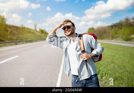 Beau jeune homme avec sac à dos, randonnée sur la route, protégeant les yeux du soleil, regardant dans la distance à la voiture qui passe Banque D'Images