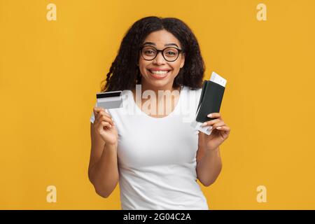 Acheter des billets d'avion en ligne. Bonne dame noire tenant passeport, billets d'avion et carte de crédit, fond jaune Banque D'Images