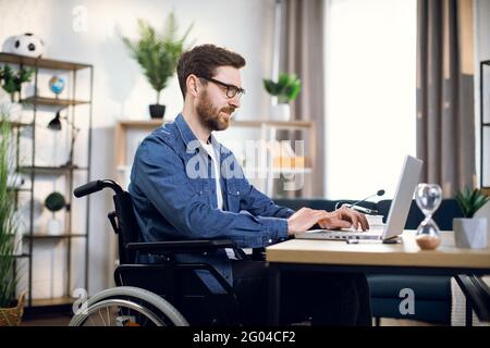 Vue latérale du jeune barbu assis en fauteuil roulant et dactylographiant sur un ordinateur portable sans fil. Beau homme handicapé dans les lunettes de travail de la maison. Banque D'Images