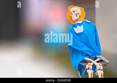 Squelette anatomique humain flexible souriant pour l'enseignement de l'anatomie médicale assis sur une chaise pliante et portant un chapeau jaune et des vêtements bleus contre un blu Banque D'Images