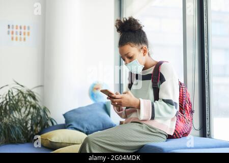 Portrait de l'adolescente afro-américaine portant un masque dans la classe scolaire et regardant un smartphone, des mesures de sécurité covid Banque D'Images