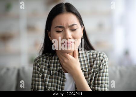 Triste femme asiatique se frottant la gorge, à l'intérieur de la maison Banque D'Images
