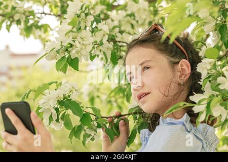 Jolie petite fille souriante utilise un smartphone pour diffuser des contenus sur les réseaux sociaux. Photo de printemps sur le fond des pommiers en fleurs Banque D'Images