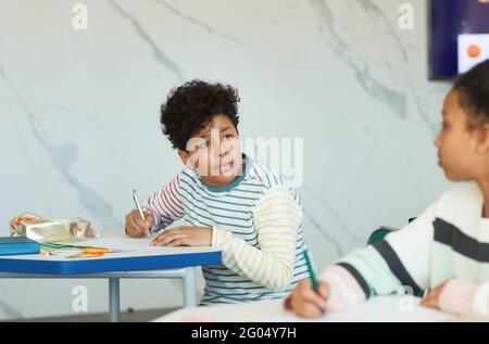 Portrait d'un jeune garçon parlant à un ami tout en étant assis à un bureau dans une salle de classe scolaire, espace de copie Banque D'Images