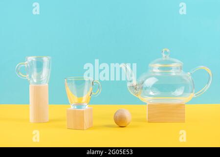 Ensemble de thé transparent en verre vide. Théière et deux tasses debout sur des formes géométriques en bois, fond jaune et bleu. Vue avant. Banque D'Images