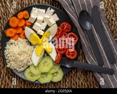 Assiette foncée sur un tapis de paille avec du riz ordinaire, des œufs durs hachés, des tomates cerises, des tranches de concombres, des carottes sur roues et des cubes de fromage grec feta Banque D'Images