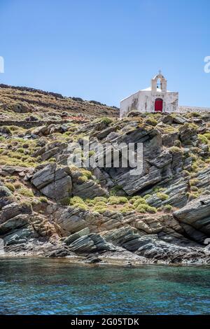 Petite église dans l'île de Kythnos, Cyclades, Grèce. Murs blanchis blancs chapelle orthodoxe avec beffroi et porte rouge sur une falaise rocheuse au-dessus de bleu clair s. Banque D'Images