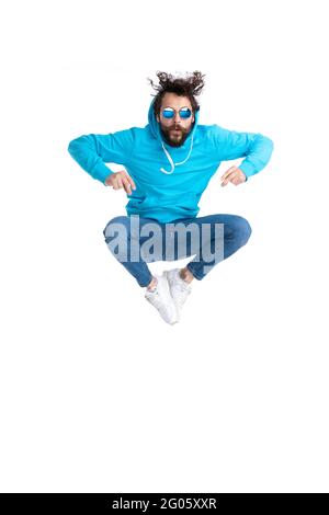 homme décontracté barbu à capuche portant des lunettes de soleil tout en se prenant les doigts et en sautant dans l'air sur fond blanc en studio Banque D'Images