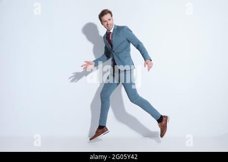 élégant jeune homme d'affaires en costume bleu regardant à côté, bondissant dans les airs tout en gestimant et en s'amusant sur fond gris en studio Banque D'Images