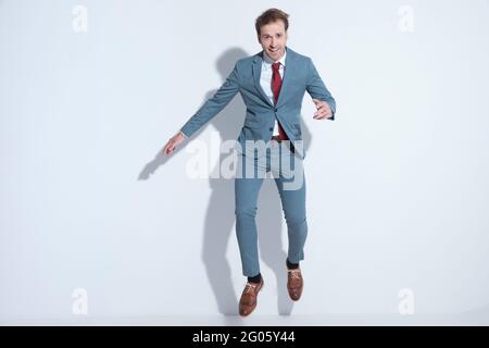 image du corps entier d'un jeune homme d'affaires enthousiaste en costume bleu élégant sautant dans les airs et s'amusant en posant sur fond gris dans stu Banque D'Images