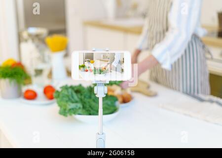 Gros plan de l'écran de smartphone avec les mains de la femme Blogger alimentaire préparant des légumes pour la salade fraîche saine tout en cuisinant dans la cuisine à la maison, enregistrement vidéo recette pour le blog. Mise au point sur l'affichage Banque D'Images