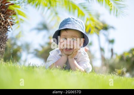 Un petit garçon qui pose sur l'herbe verte sur la pelouse sous les palmiers. Ciel bleu clair sur l'arrière-plan. Concept de vacances et d'enfance heureuse Banque D'Images