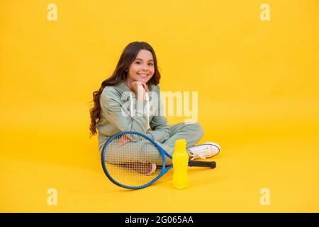 un enfant joyeux s'assoit dans des vêtements de sport avec raquette de badminton et bouteille d'eau sur fond jaune, détendez-vous Banque D'Images
