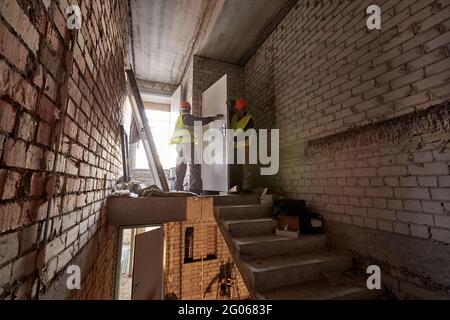 Deux ouvriers en vêtements de travail et en casques de sécurité installent une porte dans un escalier non fini d'un bâtiment en construction Banque D'Images