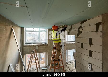 Travailleur professionnel debout sur une échelle et effectuant des mesures à l'aide d'un mètre ruban pour installer un plafond à cloisons sèches dans un bâtiment en hauteur en construction Banque D'Images