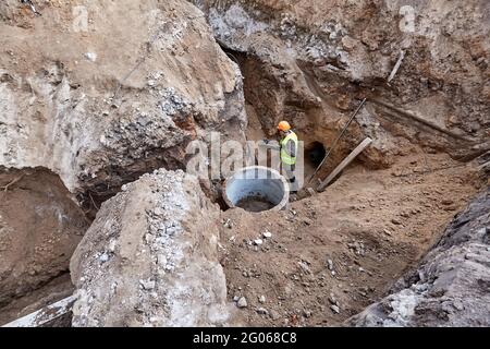 Le travailleur creuse une tranchée à l'aide d'une pelle dans une fosse d'excavation pour installer des communications dans un nouveau bâtiment en hauteur en cours de construction Banque D'Images