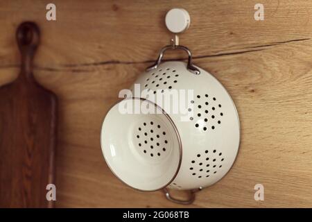 Passoire blanche en métal pendue sur un crochet sur un mur en bois de cuisine. Banque D'Images