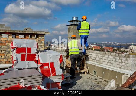 Deux hommes constructeurs professionnels de vêtements de travail et de casques orange travaillent avec un système de ventilation sur le toit du bâtiment en construction Banque D'Images