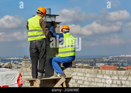 Deux hommes constructeurs professionnels de vêtements de travail et de casques de chantier installent un tuyau pour système de ventilation sur le toit du bâtiment en construction Banque D'Images