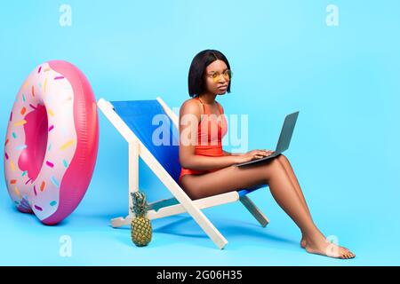 Travail en ligne pendant les vacances d'été. Une dame noire malheureuse assise dans une chaise longue avec ordinateur portable, ayant à faire du travail pendant les vacances Banque D'Images