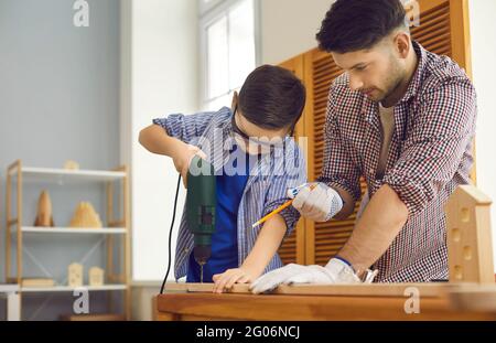 Un petit garçon sous la supervision d'un père fore une planche en bois dans l'atelier à la maison. Banque D'Images