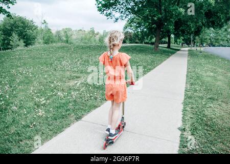 Jeune fille enfant dans la barboteuse rouge orange équitation scooter sur la route de la rue parc à l'extérieur. Été amusement ECO sport activité pour les enfants. Vue de l'arrière de behi Banque D'Images