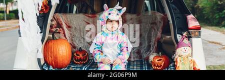 Trick ou tronc. Bébé triste bouleversé en costume de licorne célébrant Halloween dans le coffre de la voiture. Adorable tout-petit fêtant ses vacances d'octobre en plein air. Banque D'Images