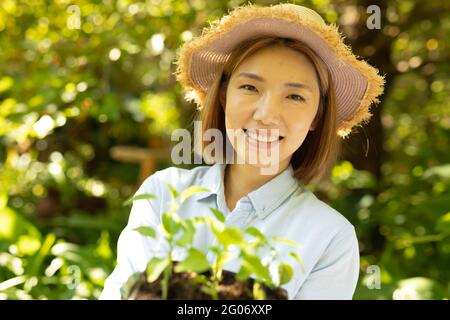 Femme asiatique souriante portant un chapeau de paille et tenant une plante dans le jardin Banque D'Images