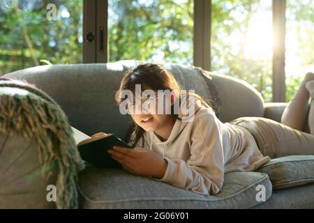 Fille asiatique souriante en lunettes lisant un livre et allongé sur un canapé Banque D'Images