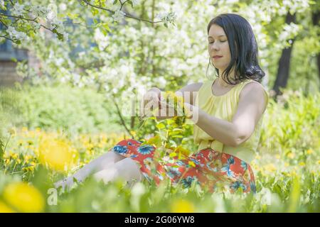 une jeune fille brune dans une robe d'été s'assoit sur une pelouse verte et tisse une couronne de fleurs de pissenlit. Banque D'Images