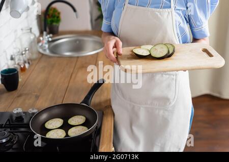 vue rognée d'une jeune femme adulte tenant des tranches d'aubergine sur le plan de coupe près d'une poêle dans la cuisine Banque D'Images