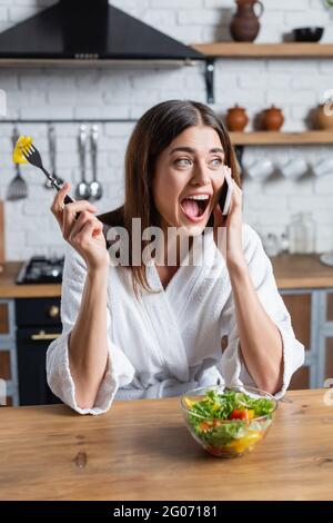 jeune femme adulte en peignoir de manger de la salade de légumes et de parler au téléphone portable dans la cuisine moderne Banque D'Images