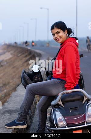 Une jolie femme indienne vêtue de rouge à cheval sur un scooter noir à côté de la route et souriant à la caméra Banque D'Images