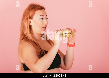 Une femme affamée de chubby tient un hamburger de fast food dans ses mains sur le point du manger, debout sur fond rose, portrait de studio. Banque D'Images