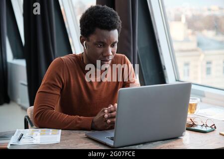 Un jeune homme d'affaires africain dans des écouteurs assis à la table et regardant le moniteur de l'ordinateur tout en étant assis au bureau Banque D'Images