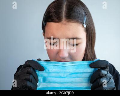 Une adolescente en gants en latex noir tenant et éternuant dans un masque médical de protection bleu. Yeux fermés. Concept de protection antivirale. Banque D'Images