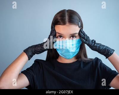 Une adolescente dans un masque médical de protection bleu touchant ses temples avec les mains dans des gants en latex noir. Concept d'infection virale. Banque D'Images