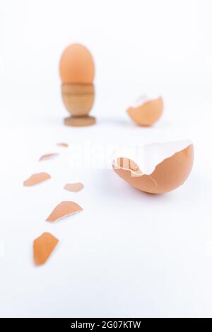 Tirer verticalement l'œuf de poule dans un ancien support d'œufs en bois et plusieurs coquilles d'œufs cassées sur une surface blanche. Minimalisme. Art conceptuel pour l'alimentation ou le produit animal Banque D'Images